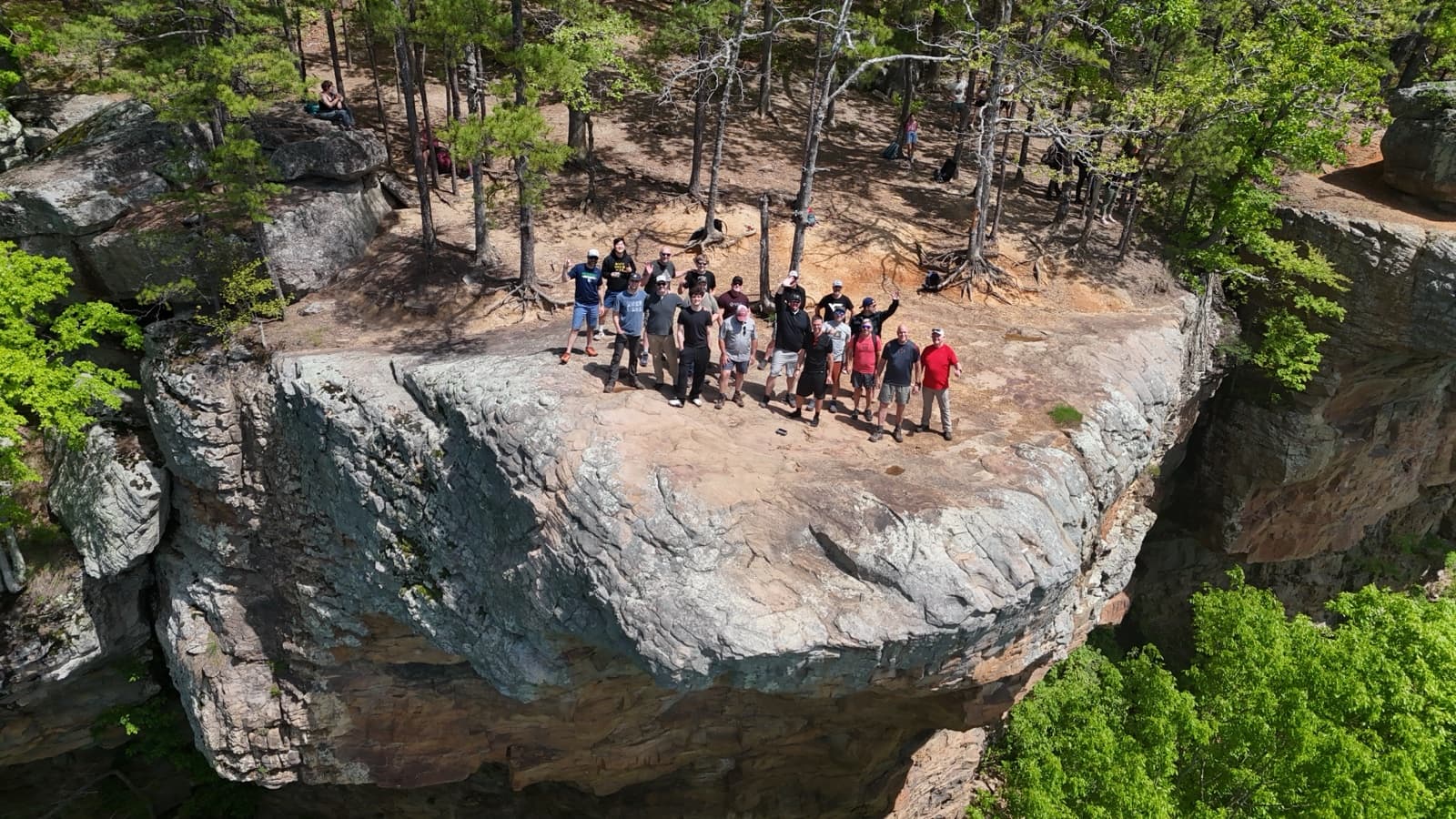 Drone shot of a hiking group waving from a huge sandstone bluff in the Ozarks, pines all around.