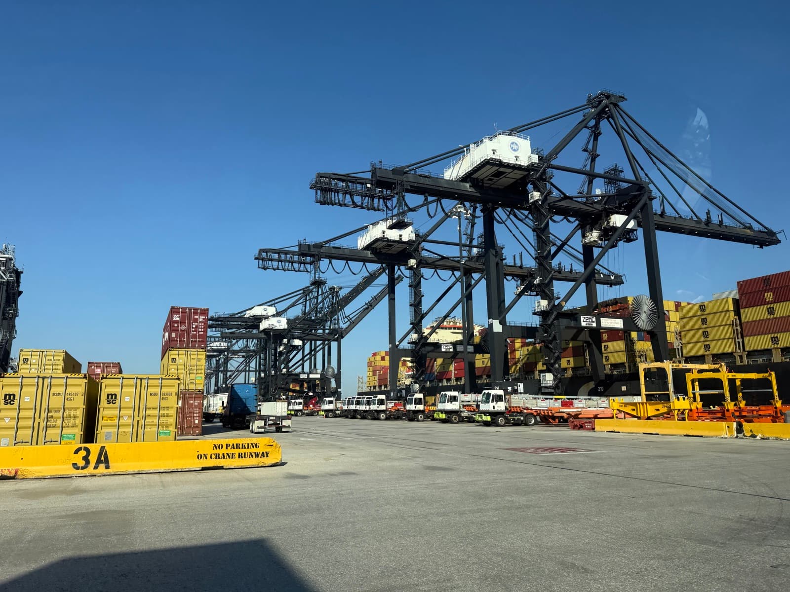 Container cranes towering over stacked shipping containers at a port under a clear sky.