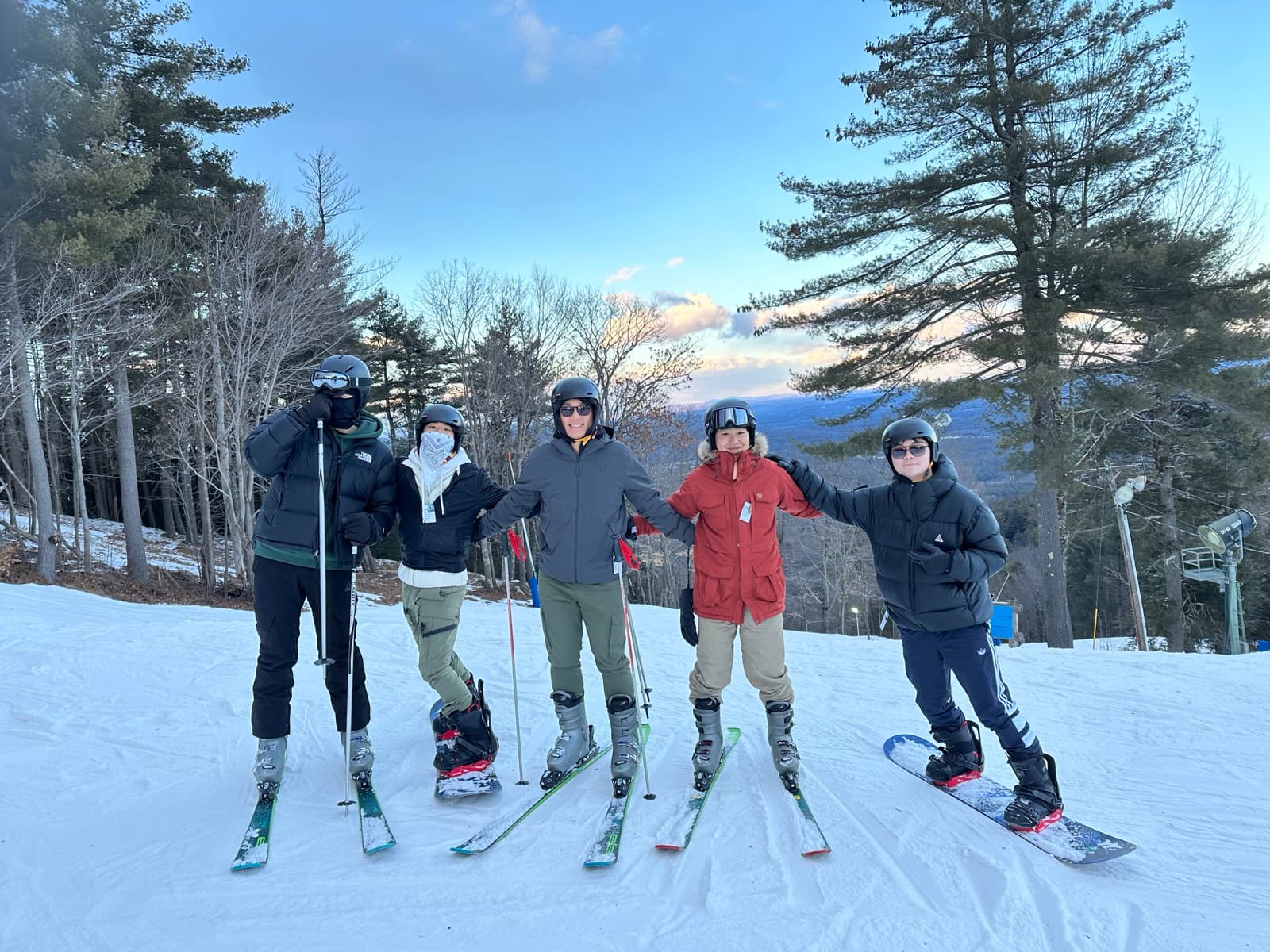 Five friends in ski gear lined up on the slope at dusk, mountains behind.