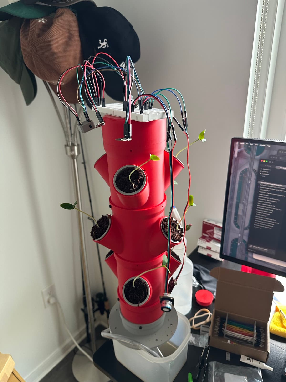 A red hydroponic grow tower mid-build, sensor wires sprouting from the top like a second crop.