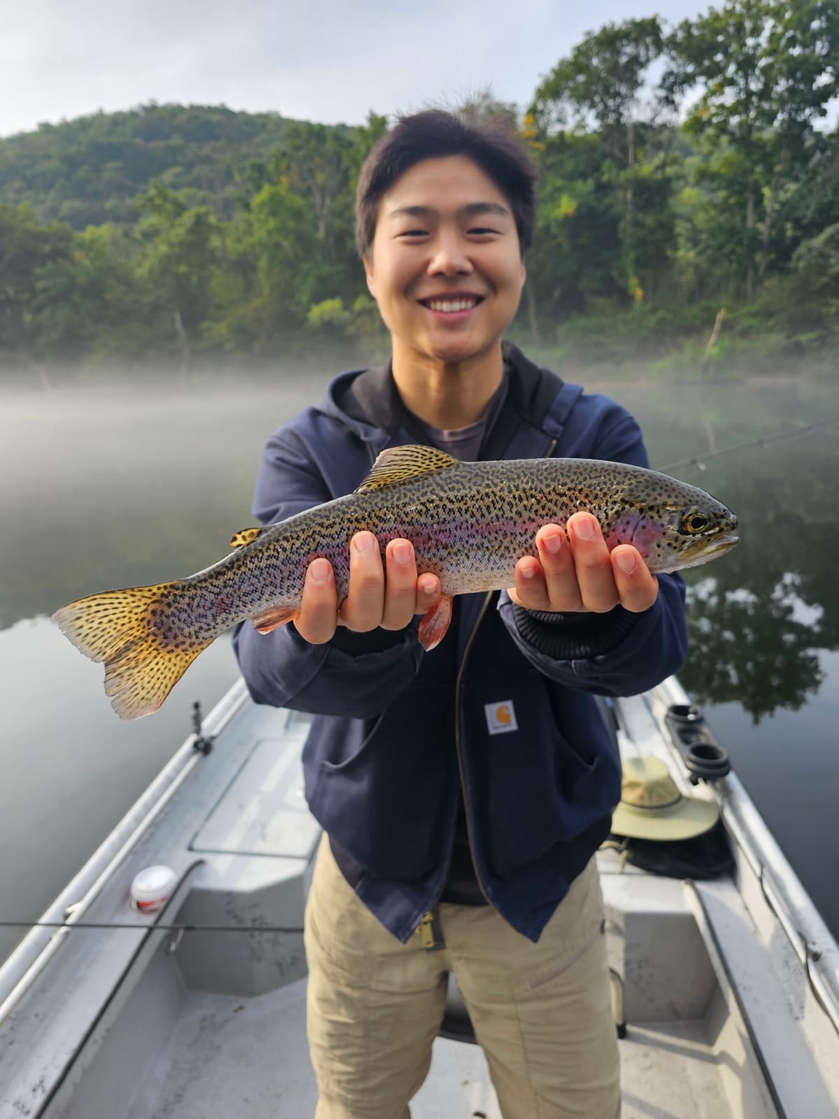 Paul on a drift boat holding a rainbow trout, morning fog still on the water.