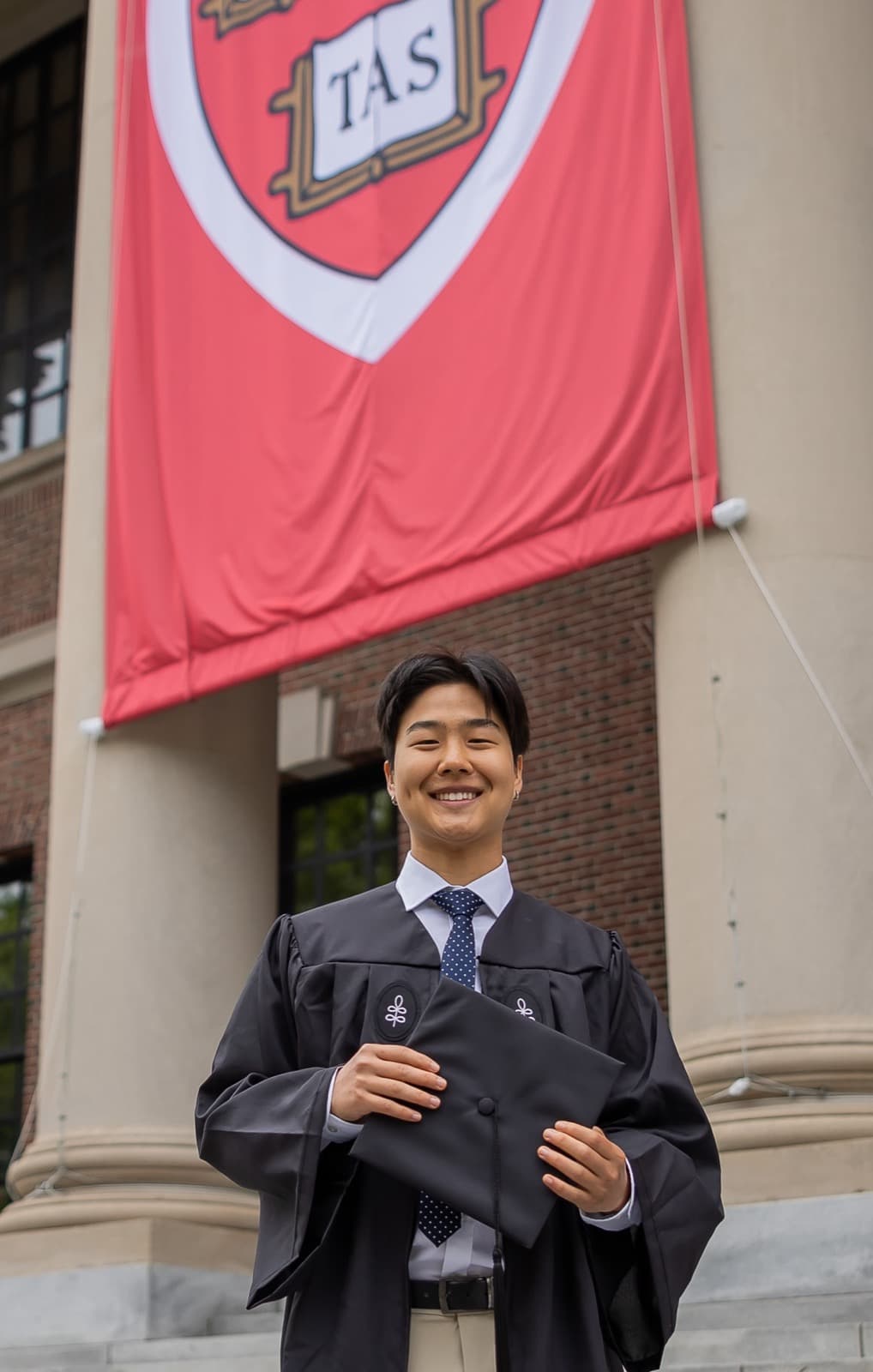 Paul Song at Harvard commencement — smiling in cap and gown in front of a crimson Veritas banner.