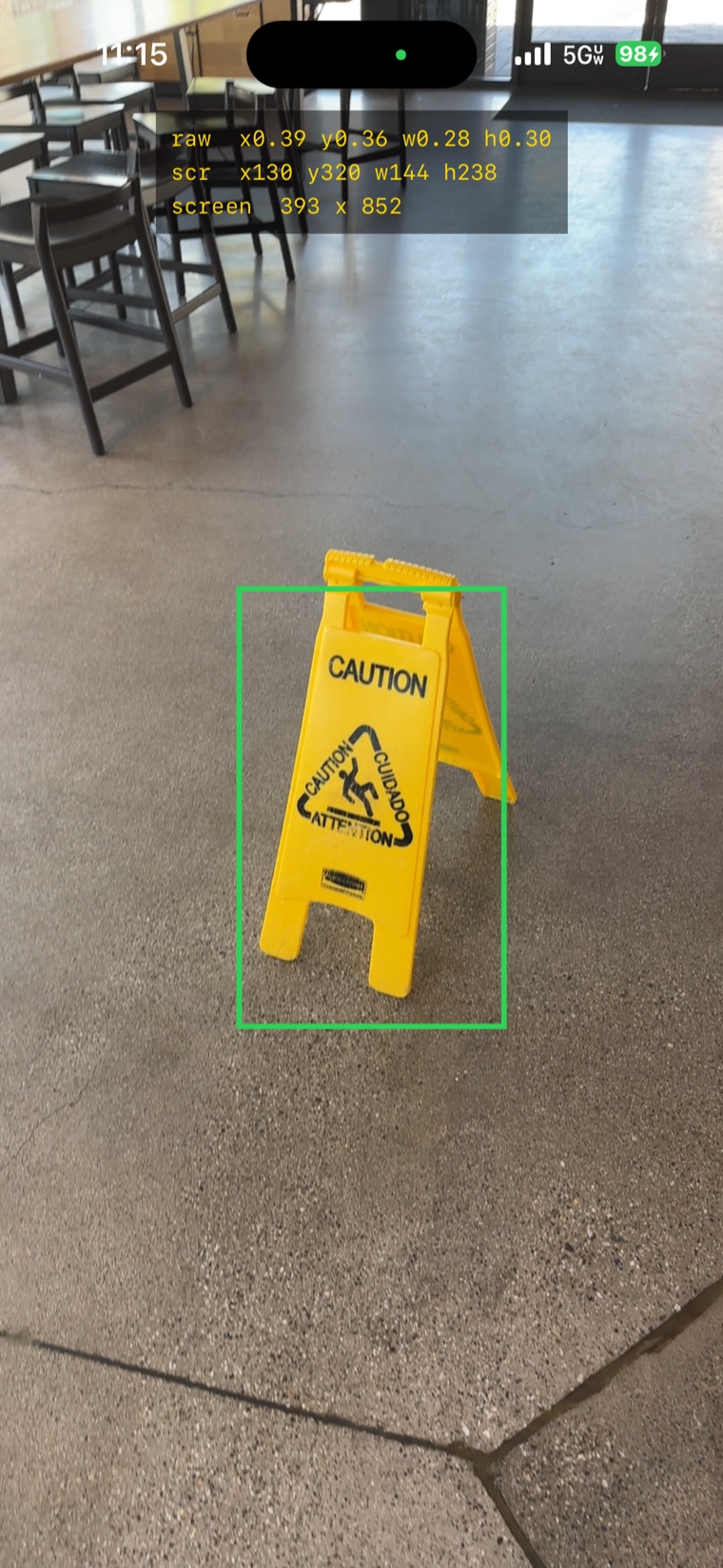 A phone screenshot of the app: a green bounding box snapped tightly around a yellow CAUTION wet-floor sign, with the debug HUD overlaid at the top showing the raw normalized rect, the converted screen-pixel rect, and the screen size.